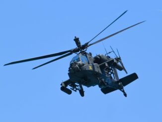 Američki jurišni helikopter AH‑64 Apache u letu, snimljen iz blizine u niskom preletu iznad terena, s jasno vidljivim rotorskim krilima i naoružanjem.